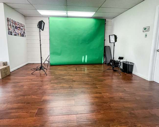 The image shows a room with a green screen backdrop, lighting equipment, and wooden flooring.