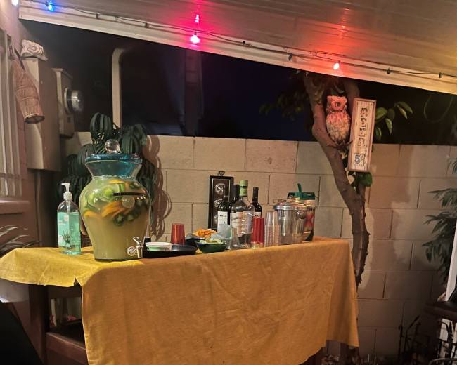 A wooden bar with a yellow tablecloth is set up outdoors, featuring cocktail glasses and a large pitcher filled with a drink.