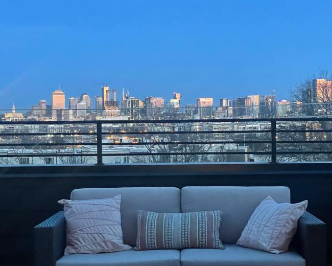 A balcony features a sofa facing a city skyline illuminated at dusk.