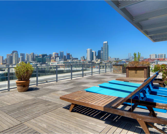 The image shows a rooftop terrace featuring wooden decking, lounge chairs, and a view of a city skyline under a clear blue sky.