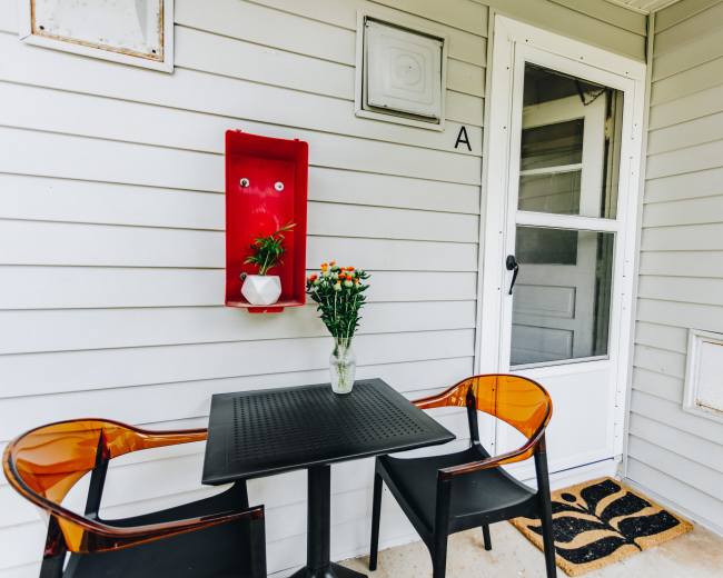 A small patio area features a black table with two transparent chairs, a flower vase, and a door leading inside.