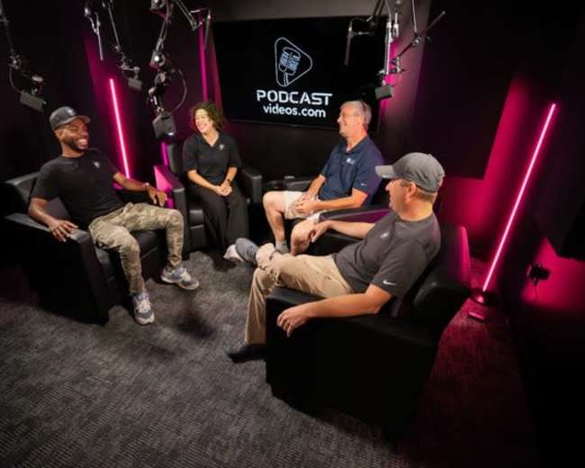 A group of four individuals sits in chairs in a podcast studio, surrounded by camera equipment and soft lighting.