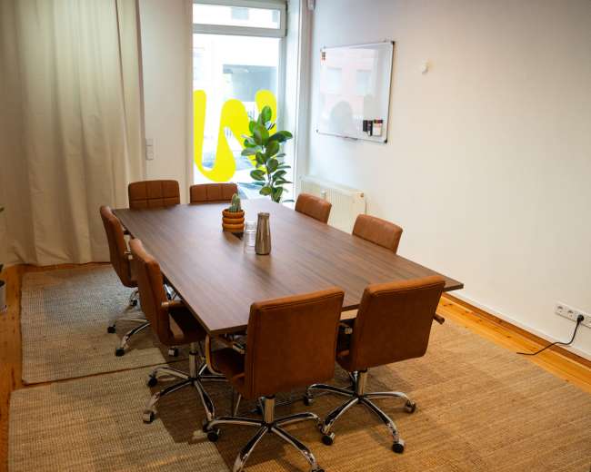 The image shows a modern conference room with a large wooden table surrounded by brown leather chairs, a potted plant in the corner, and natural light streaming in through a window.