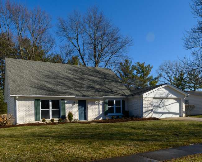 A single-story house with a gabled roof, green shutters, and a two-car garage is situated on a lawn surrounded by trees.