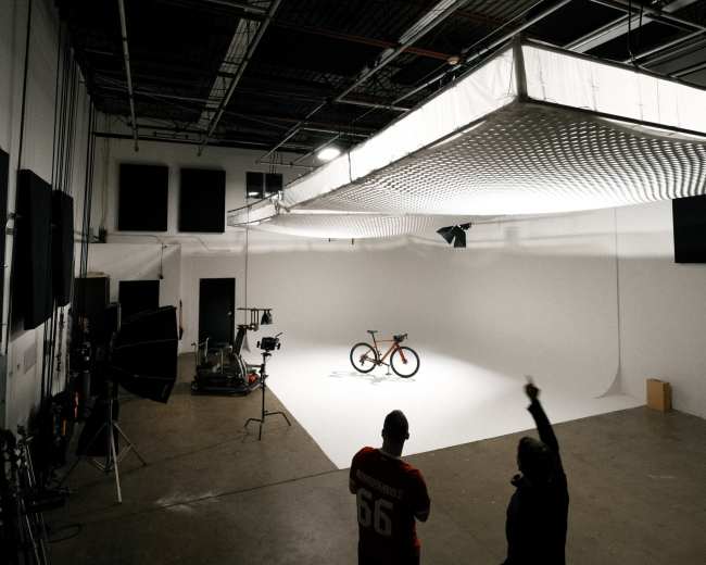 Two people stand in a photo studio with a bicycle on a white backdrop under bright lights.
