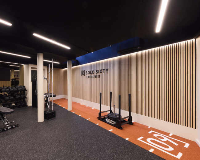 The image shows a modern gym interior with weightlifting equipment, a sled, and a designated exercise area featuring a turf surface.