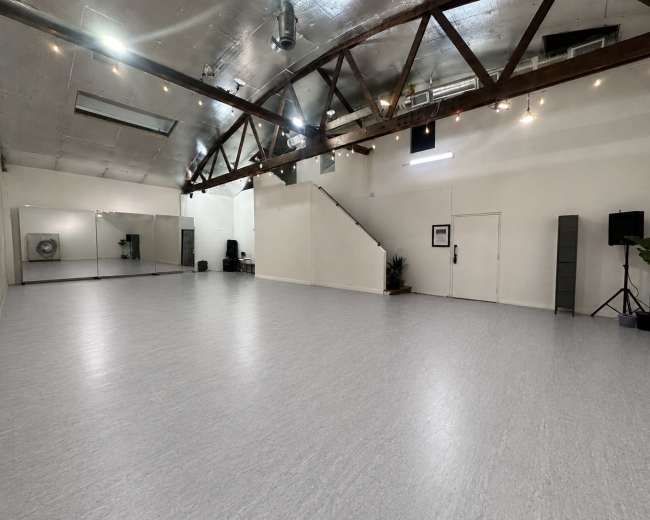 The image shows a spacious, well-lit dance studio with exposed wooden beams, mirrors on one wall, and a staircase in the background.