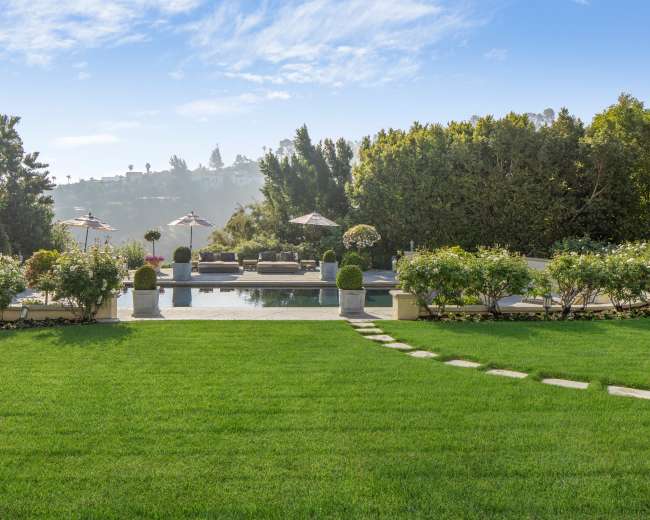The image shows a lush green lawn leading to a tranquil pool area surrounded by landscaping and outdoor seating under umbrellas.