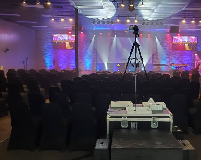 A spacious auditorium is set up with rows of black chairs facing a stage equipped with lighting and a camera on a tripod, while two screens display visuals at the front.
