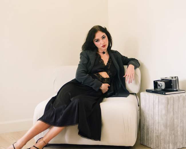 A woman in a black dress and blazer sits on a light-colored sofa, with a vintage camera on a nearby table.