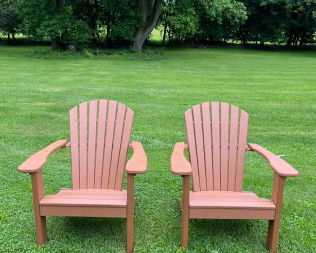 Two Adirondack chairs are positioned side by side on a grassy lawn surrounded by trees.