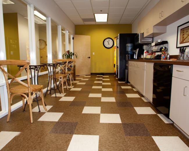 The image shows a break room with a tiled floor, a kitchenette along one wall, a dining table with chairs, and large windows on the opposite side.