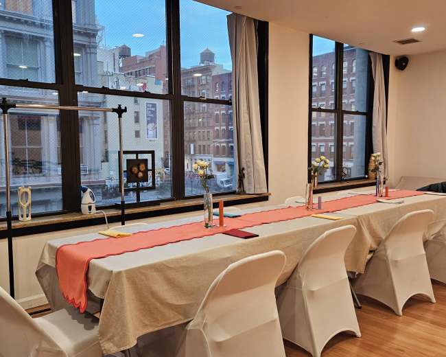 A long table set with beige tablecloths and centerpieces is arranged near large windows offering a view of a city street.