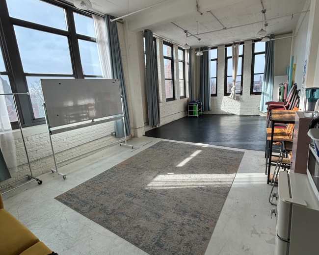 The image shows a spacious, bright room with large windows, a gray area rug on the floor, a whiteboard, and a few chairs arranged near the walls.