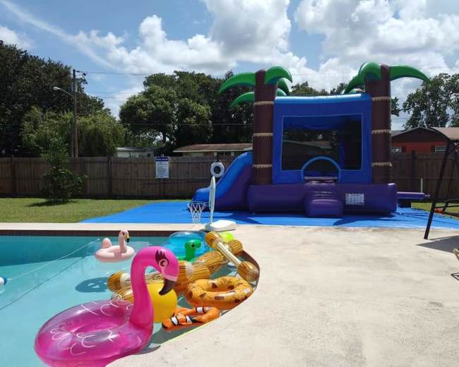 A backyard scene features an in-ground pool with colorful inflatable toys alongside a bounce house with palm tree decorations and a basketball hoop.