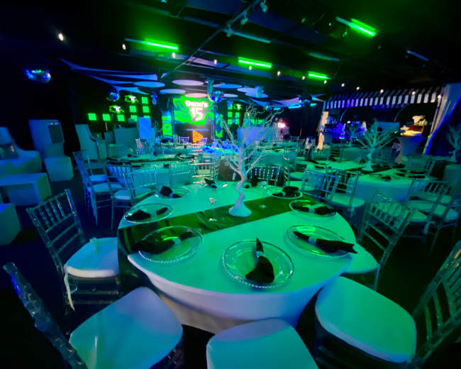 The image shows a brightly lit event venue with circular tables set for a celebration, decorated with black napkins and white centerpieces, under green and blue ambient lighting.