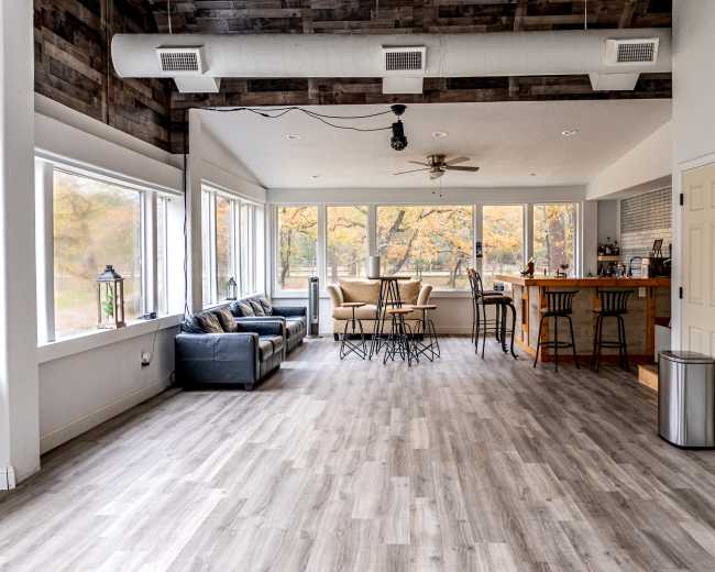 The image shows a spacious, modern living area with large windows, a seating area, a dining table, and a kitchen counter.