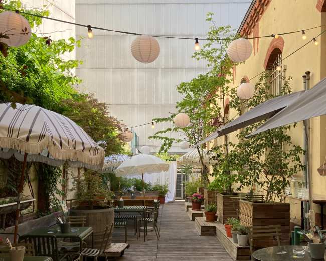 A narrow wooden walkway leads through a garden area filled with potted plants and tables, adorned with hanging lanterns and umbrellas.