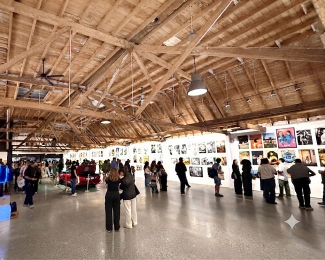 A crowd of people observes a gallery exhibition featuring a series of framed photographs displayed on the walls of a spacious, wooden-beamed venue.
