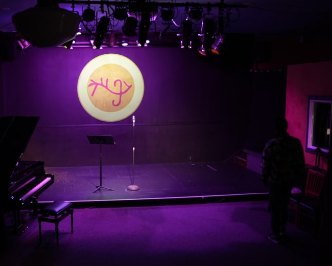 An empty performance stage with a grand piano on the left, a microphone stand centered, and a circular logo illuminated on the back wall.