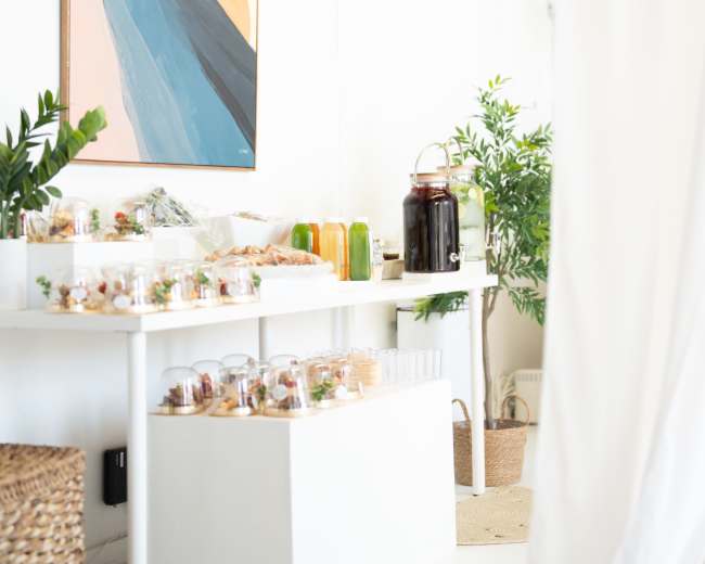 A minimalist white table displays an array of food items and drinks, set against a large abstract painting on the wall.