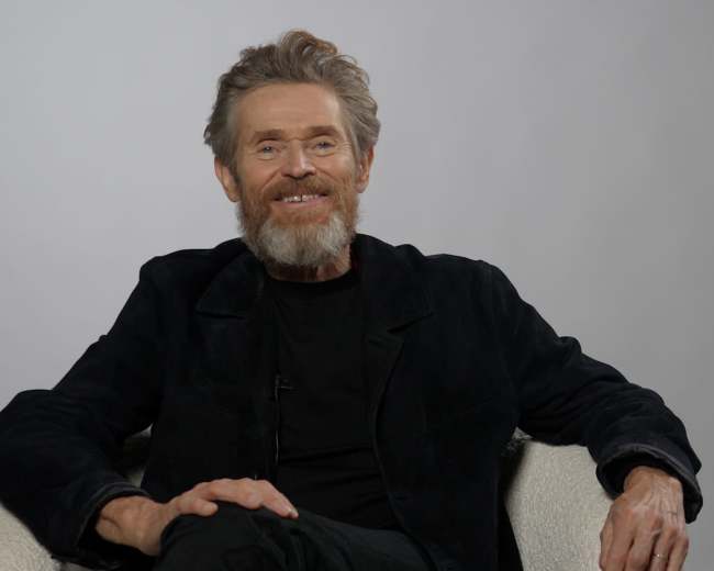 A man with a beard is sitting comfortably in a chair against a plain light background.