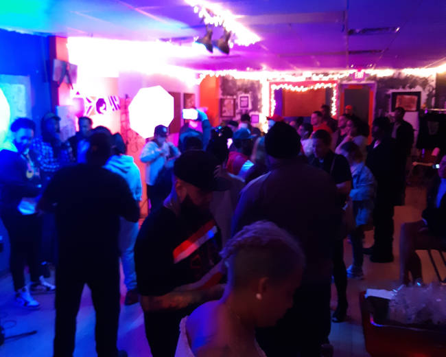 A crowded room filled with people socializing under colorful lights, with some individuals holding up their phones.