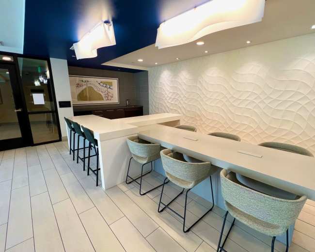 A modern kitchen or bar area with a long, white countertop and high-backed chairs against a textured wall.