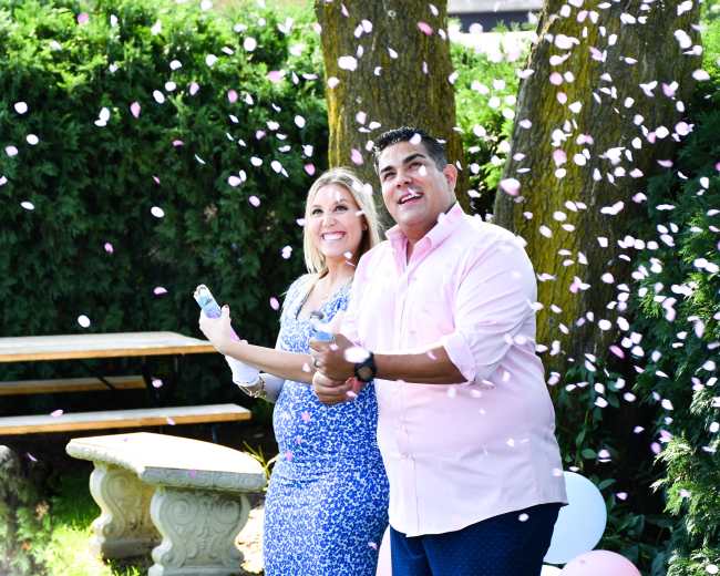A couple stands together in a garden, smiling as pink confetti falls around them, celebrating a joyful moment.