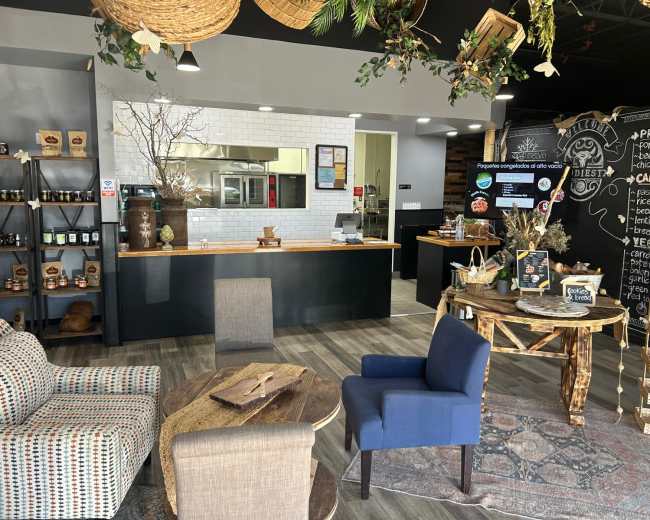 The image depicts a cozy café interior featuring a kitchen area, a seating arrangement with armchairs, and a chalkboard menu.