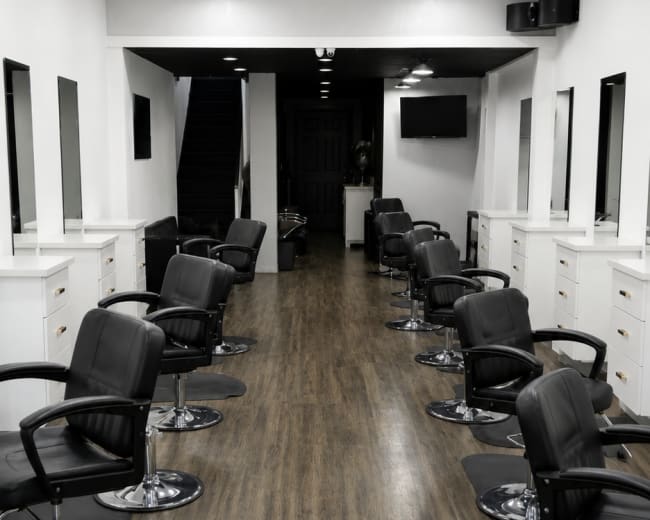 The image shows a modern hair salon with several black salon chairs arranged along a wooden floor and large mirrors on the walls.