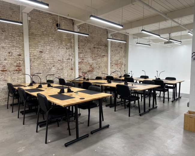 The image shows a spacious room with several wooden desks and black chairs arranged in rows against a brick wall.