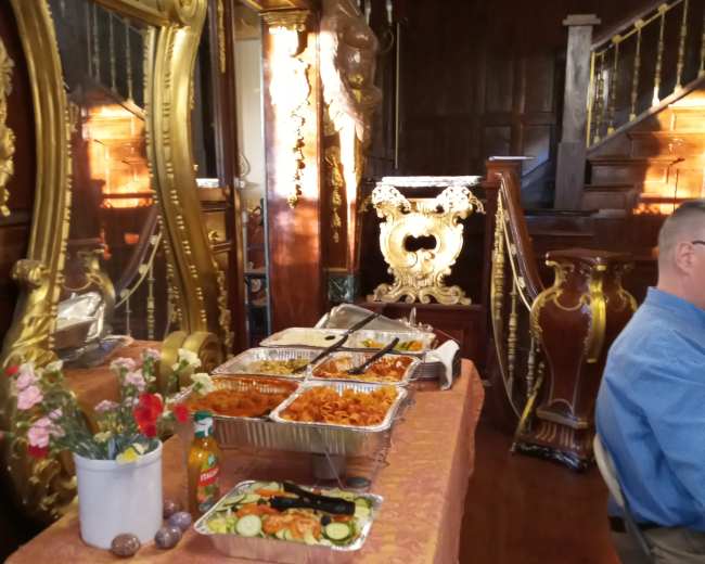 A table covered with a patterned cloth holds several trays of food, accompanied by a vase of flowers, in an ornate room featuring large mirrors and decorative wooden elements.
