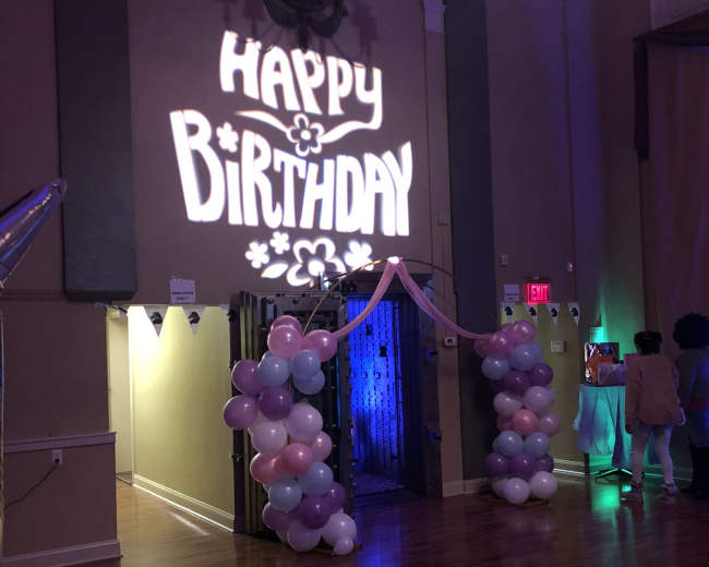 The scene features a birthday party setting with a "Happy Birthday" projection on the wall and an entrance decorated with pink and blue balloons.