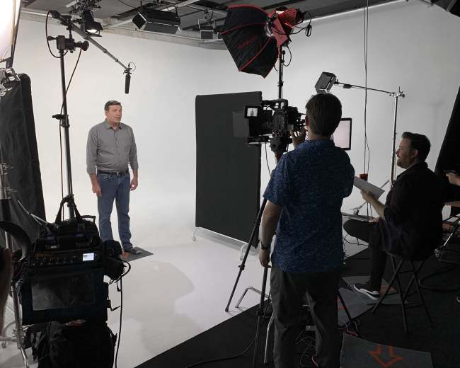 A man stands in front of a camera setup on a simple studio set, while a crew operates equipment in the background.