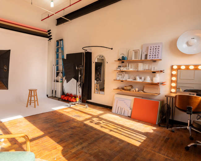 A photography studio with a white backdrop, lighting equipment, a stool, and organized shelves displaying various items.