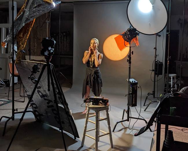 A model stands on a stool surrounded by photography equipment and backdrops in a studio setting.