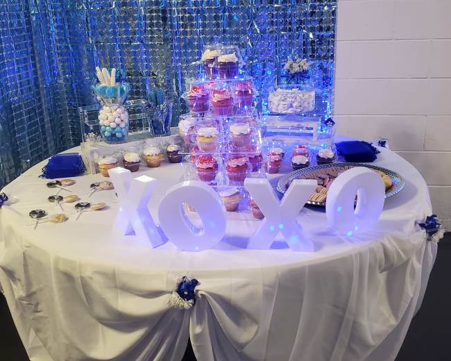 A table is set with a blue and white tablecloth featuring a tiered cupcake display, assorted sweets, and illuminated "XOXO" letters in front of a shimmering backdrop.