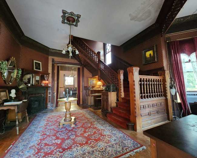 The image features a vintage foyer with a large staircase, ornate woodwork, and a decorative rug on a wooden floor.