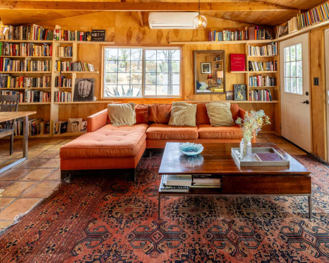 The image shows a cozy interior of a room featuring a large orange sectional sofa, a wooden coffee table, bookshelves lined with books, and a door leading outside.
