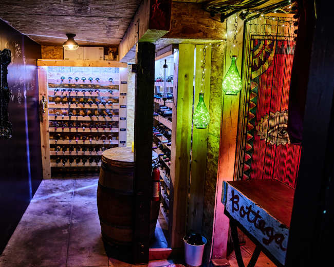 A dimly lit wine cellar with wooden shelves filled with bottles, a barrel, and decorative lighting.