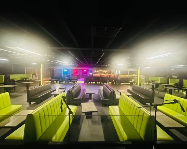 The image shows an empty indoor venue with green upholstered seating, dim lighting, and a bar area in the background.