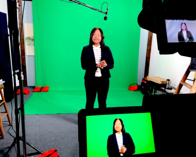 A woman stands in front of a green screen set, with cameras and equipment positioned to capture her presentation.