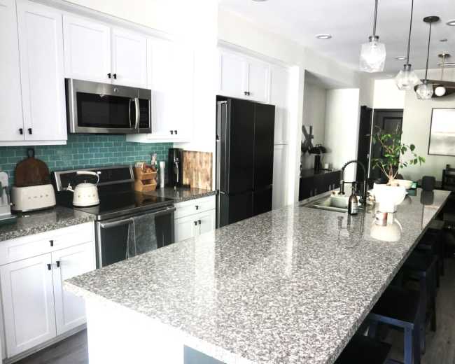 The image shows a modern kitchen with white cabinetry, stainless steel appliances, a granite countertop, and a dining area in the background.