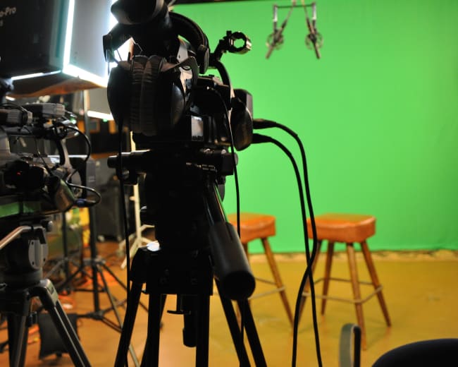A camera setup is positioned in front of two empty stools against a green screen backdrop in a studio setting.