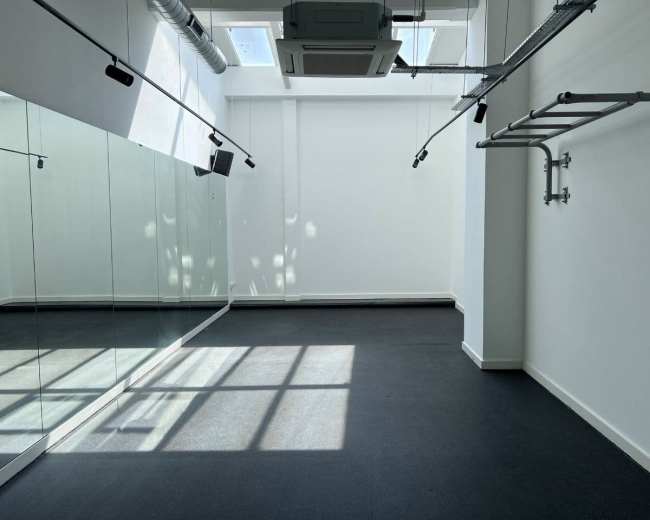 The image shows a spacious room with mirrored walls, a ceiling-mounted air conditioning unit, and a section of exposed piping, illuminated by natural light from overhead windows.