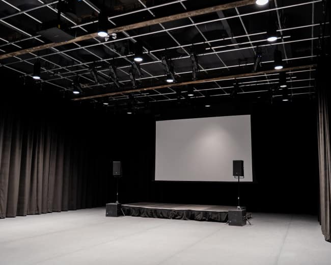 The image shows an empty stage with a large screen and audio equipment in a dimly lit auditorium space.