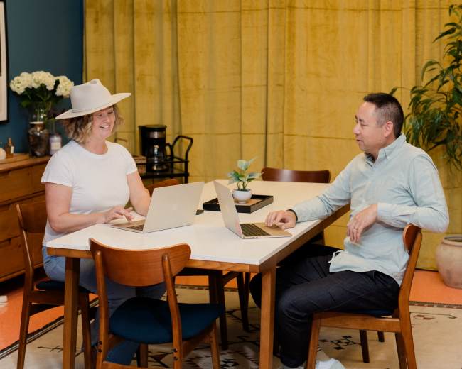 Two people are sitting at a table with laptops in a room featuring blue walls and yellow curtains.
