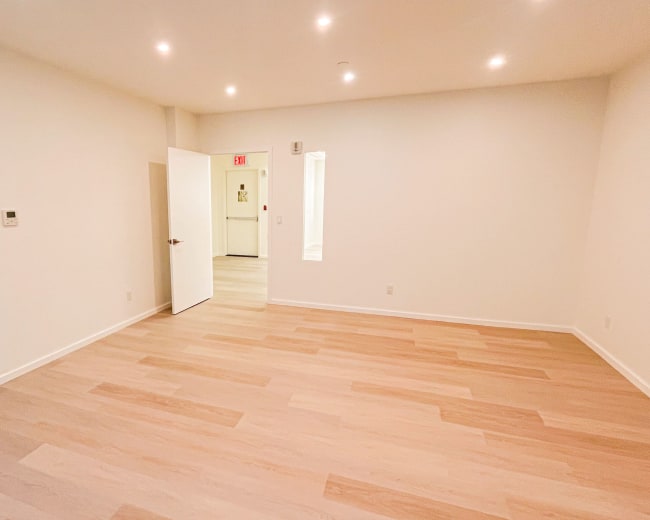 The image shows a sparsely furnished room with light wood flooring, white walls, and recessed lighting.
