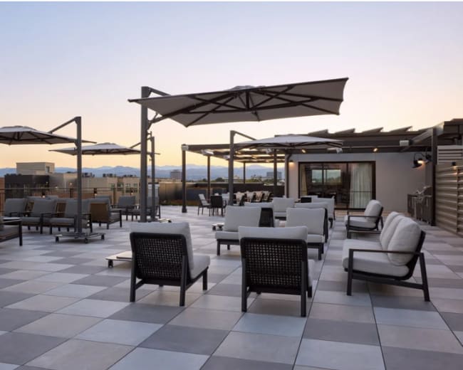 The image shows a rooftop terrace with gray and white tiled flooring, featuring several lounge chairs and tables under large umbrellas, with mountains visible in the background.